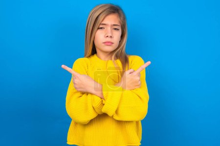 Photo for Serious beautiful blonde teen girl wearing yellow sweater over blue wall crosses hands and points at different sides hesitates between two items. Hard decision concept - Royalty Free Image