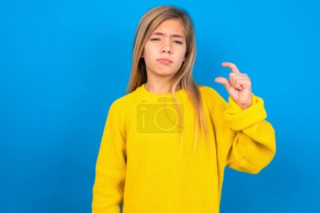 Téléchargez les photos : Mauvaise belle adolescente blonde portant un pull jaune sur des formes de mur bleu petit signe de la main démontre quelque chose de pas très grand. Concept de langage corporel. - en image libre de droit