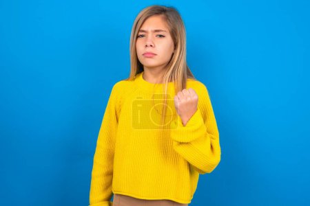 Photo for Beautiful blonde teen girl wearing yellow sweater over blue wall shows fist has annoyed face expression going to revenge or threaten someone makes serious look. I will show you who is boss - Royalty Free Image