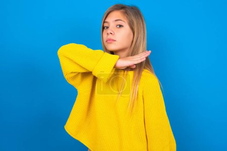 Photo for Beautiful blonde teen girl wearing yellow sweater over blue wall cutting throat with hand as knife, threaten aggression with furious violence. - Royalty Free Image