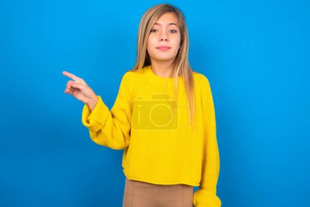 Foto de Positivo hermosa rubia adolescente chica usando suéter amarillo sobre la pared azul con expresión satisfecha indica en la esquina superior derecha muestra buena oferta sugiere hacer clic en el enlace - Imagen libre de derechos