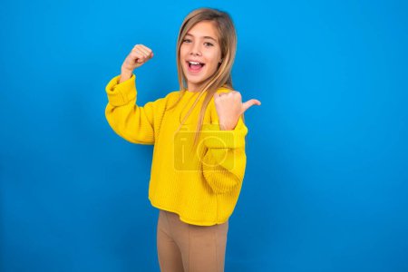 Photo for Hooray cool beautiful blonde teen girl wearing yellow sweater over blue wall point back empty space hand fist - Royalty Free Image