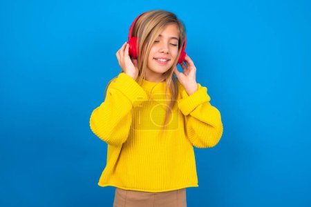 Photo for Pleased beautiful blonde teen girl wearing yellow sweater over blue wall enjoys listening pleasant melody keeps hands on stereo headphones closes eyes. Spending free time with music - Royalty Free Image