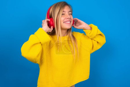 Téléchargez les photos : Belle adolescente blonde portant un pull jaune sur un mur bleu avec un casque sur la tête, écoutant de la musique, appréciant la chanson préférée avec les yeux fermés, tenant la main sur un casque. - en image libre de droit