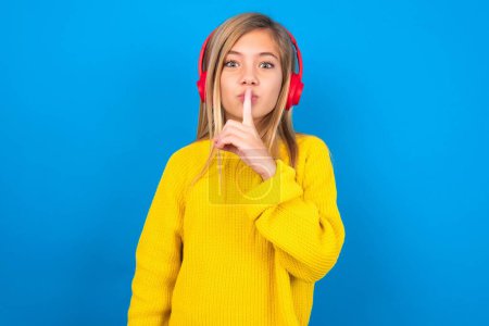 Photo for Beautiful blonde teen girl wearing yellow sweater over blue wall making hush gesture with finger on her lips wearing  wireless headphones. Be quiet. - Royalty Free Image