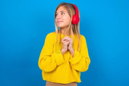 Foto de Hermosa chica adolescente rubia con suéter amarillo sobre la pared azul lleva auriculares estéreo escuchando música concentrada y mirando a un lado con interés. - Imagen libre de derechos