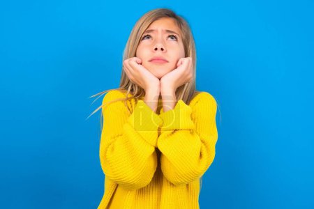 Photo for Portrait of sad beautiful blonde teen girl wearing yellow sweater over blue wall hands face look empty space - Royalty Free Image