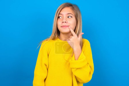 Photo for Lovely dreamy beautiful blonde teen girl wearing yellow sweater over blue wall keeps finger near lips looks aside copy space. - Royalty Free Image