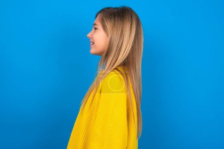 Foto de Perfil retrato de agradable hermosa rubia adolescente chica usando suéter amarillo sobre la pared azul mirada espacio vacío sonrisa dentada - Imagen libre de derechos