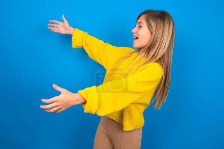 Foto de Divertido asombrado hermosa rubia adolescente chica usando suéter amarillo sobre la pared azul mirada espacio vacío con los brazos abiertos listo para coger algo. - Imagen libre de derechos