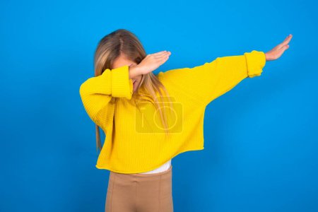 Photo for Photo of funky beautiful blonde teen girl wearing yellow sweater over blue wall show disco move dab - Royalty Free Image