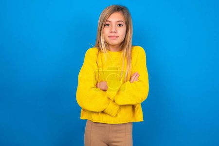 Foto de Confiado hermosa rubia adolescente chica usando suéter amarillo sobre la pared azul con los brazos cruzados mirando a la cámara - Imagen libre de derechos