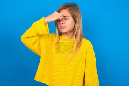 Foto de Triste hermosa rubia adolescente chica usando amarillo suéter sobre azul pared sufrimiento de dolor de cabeza celebración de la mano en su cara - Imagen libre de derechos
