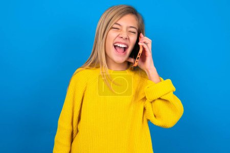 Foto de Overemotive feliz hermosa rubia adolescente chica usando suéter amarillo sobre la pared azul se ríe positivamente escucha historia divertida de amigo durante la conversación telefónica - Imagen libre de derechos