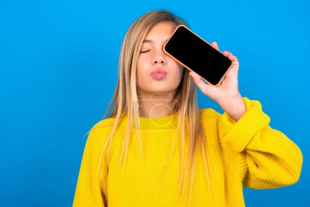 Foto de Adorable hermosa rubia adolescente chica usando suéter amarillo sobre la pared azul sosteniendo dispositivo moderno que cubre el ojo con los labios enfurecidos - Imagen libre de derechos