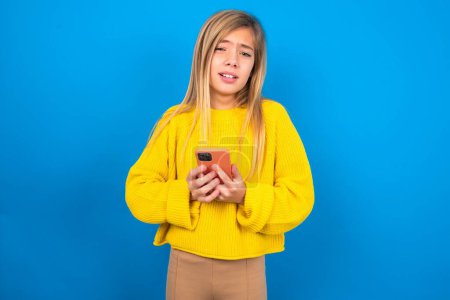 Foto de Foto de asombrado loco hermosa rubia adolescente chica usando suéter amarillo sobre la pared azul mantenga teléfono inteligente concepto de retroalimentación disgusta - Imagen libre de derechos