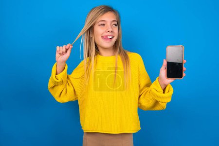 Photo for Photo of nice pretty beautiful blonde teen girl wearing yellow sweater over blue wall demonstrate phone screen hold hair tails - Royalty Free Image