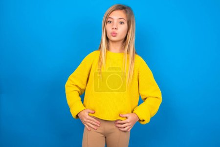 Téléchargez les photos : Portrait de belle jeune fille blonde drôle portant un pull jaune sur un mur bleu envoyant un baiser d'air - en image libre de droit