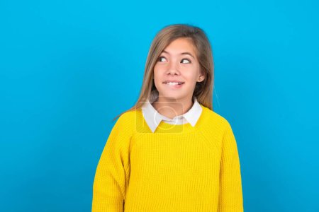 Foto de Increíble chica adolescente caucásica usando suéter amarillo sobre la pared azul mordiendo labio y buscando difícil de espacio vacío. - Imagen libre de derechos