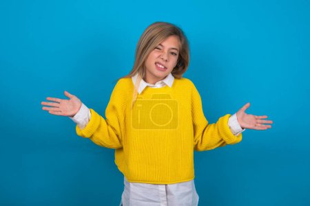 Téléchargez les photos : Clueless caucasien adolescent fille portant un pull jaune sur le mur bleu hausse les épaules avec hésitation, fait face à une situation douteuse, écarte les paumes, Décision difficile - en image libre de droit
