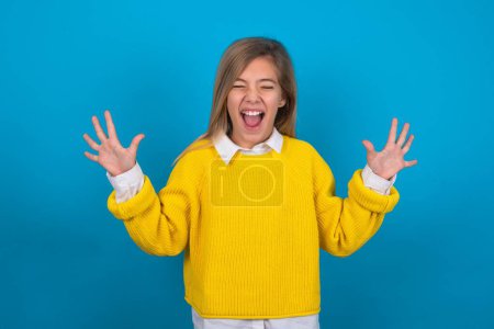 Photo for Emotive caucasian teen girl wearing yellow sweater over blue wall laughs loudly, hears funny joke or story, raises palms with satisfaction, - Royalty Free Image