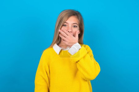 Foto de Emocional chica adolescente caucásica usando suéter amarillo sobre jadeos de pared azul de asombro, cubre la boca abierta con la palma, se ve sorprendido por la cámara. - Imagen libre de derechos