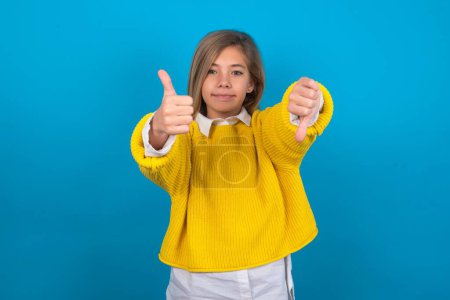 Foto de Caucásico adolescente chica usando amarillo suéter sobre azul pared mostrando pulgares hacia arriba y pulgares hacia abajo, difícil elegir concepto - Imagen libre de derechos