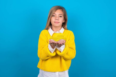Photo for Caucasian teen girl wearing yellow sweater over blue wall holding something with open palms, offering to the camera. - Royalty Free Image