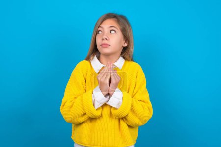 Photo for Charming cheerful caucasian teen girl wearing yellow sweater over blue wall making up plan in mind holding hands together, setting up an idea. - Royalty Free Image