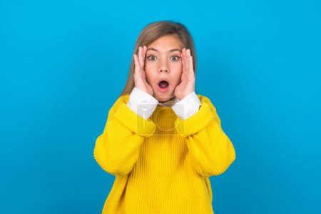 Photo for Caucasian teen girl wearing yellow sweater over blue wall with scared expression, keeps hands on head, jaw dropped, has terrific expression. Omg concept - Royalty Free Image