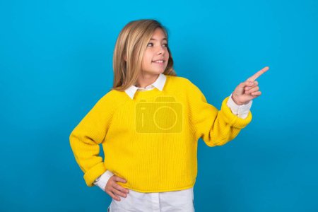 Photo for Smiling caucasian teen girl wearing yellow sweater over blue wall indicating finger empty space showing best low prices - Royalty Free Image