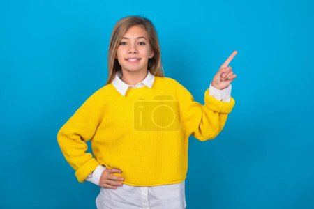 Photo for Caucasian teen girl wearing yellow sweater over blue wall looking at camera indicating finger empty space sales - Royalty Free Image