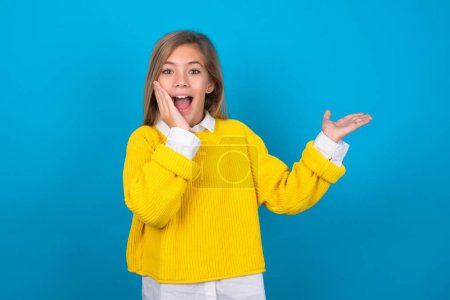 Téléchargez les photos : Fou caucasien adolescent fille portant jaune pull sur bleu mur conseiller rabais prix tenir ouvert palme nouveau produit - en image libre de droit