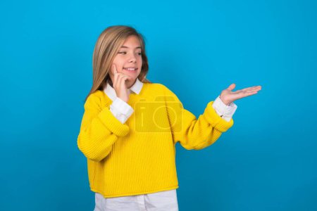 Téléchargez les photos : Funny caucasien adolescent fille portant pull jaune sur le mur bleu tenant la paume ouverte nouveau produit. Je veux l'acheter.! - en image libre de droit