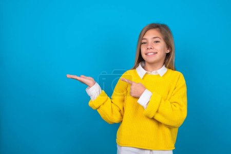 Foto de Caucásico adolescente chica usando amarillo suéter sobre azul pared apuntando y sosteniendo la mano mostrando anuncios - Imagen libre de derechos
