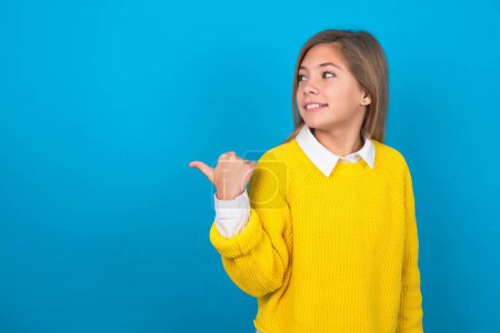 Photo for Charming caucasian teen girl wearing yellow sweater over blue wall looking at copy space having advertisements - Royalty Free Image