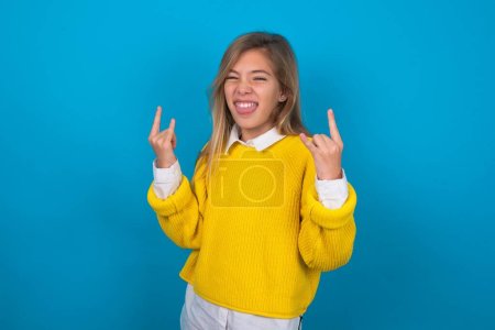Téléchargez les photos : Caucasien adolescent fille porter jaune pull sur bleu mur faisant rock main geste et montrant la langue - en image libre de droit