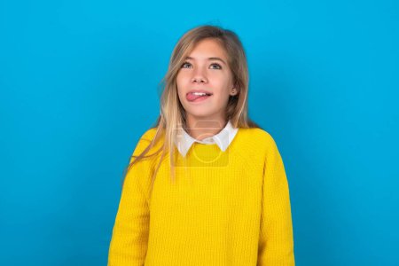 Téléchargez les photos : Adolescent caucasien fille portant un pull jaune sur le mur bleu montrant grimace visage croisant les yeux et montrant la langue. Être drôle et fou - en image libre de droit