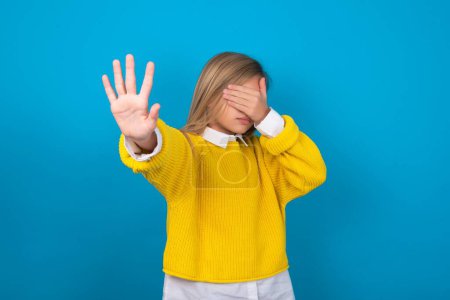 Photo for Caucasian teen girl wearing yellow sweater over blue wall covers eyes with palm and doing stop gesture, tries to hide from everybody. - Royalty Free Image