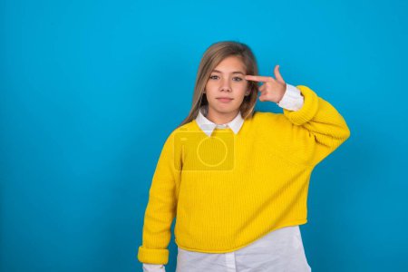 Photo for Unhappy caucasian teen girl wearing yellow sweater over blue wall makes suicide gesture and imitates gun with hand, curves lips keeps two fingers on temple, shoots, being tired of everything, - Royalty Free Image