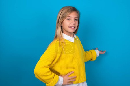 Foto de Chica adolescente caucásica vistiendo suéter amarillo sobre la pared azul sintiéndose feliz y alegre, sonriendo y dándote la bienvenida, invitándote con un gesto amistoso - Imagen libre de derechos