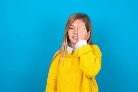 Téléchargez les photos : Caucasien adolescent fille portant pull jaune sur le mur bleu couvrant un oeil avec sa main, sourire confiant sur le visage et l'émotion surprise. - en image libre de droit