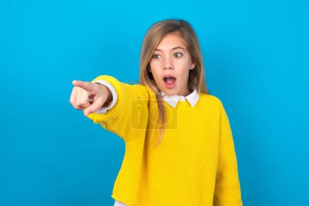 Photo for Caucasian teen girl wearing yellow sweater over blue wall Pointing with finger surprised ahead, open mouth amazed expression, something on the front. - Royalty Free Image
