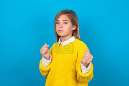 Téléchargez les photos : Caucasien adolescent fille porter jaune pull sur le mur bleu faire geste d'argent avec les mains, demander le paiement du salaire, millionnaire entreprise - en image libre de droit
