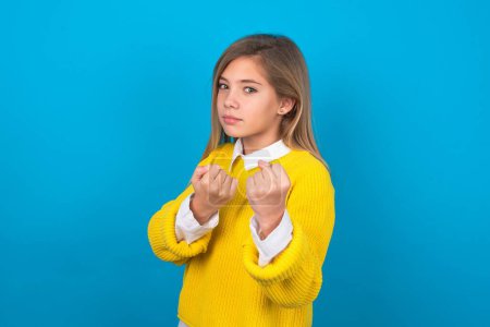 Téléchargez les photos : Caucasien adolescent fille portant pull jaune sur le mur bleu Prêt à se battre avec poing geste de défense, en colère et contrarié visage, peur de problème. - en image libre de droit