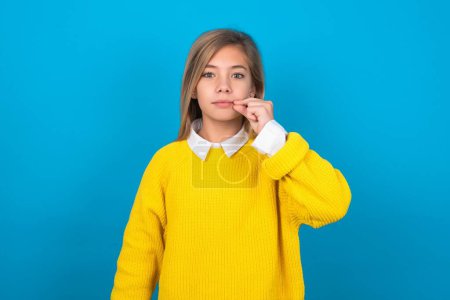 Foto de Chica adolescente caucásica con suéter amarillo sobre la boca de la pared azul y los labios cerrados como cremallera con los dedos. Secreto y silencioso, tabú hablando. - Imagen libre de derechos