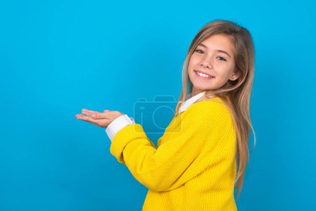 Téléchargez les photos : Caucasien adolescent fille portant jaune pull sur le mur bleu pointant de côté avec les mains ouvertes paumes montrant l'espace de copie, présentant la publicité souriant heureux excité - en image libre de droit