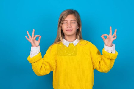 Photo for Caucasian teen girl wearing yellow sweater over blue wall relax and smiling with eyes closed doing meditation gesture with fingers. Yoga concept. - Royalty Free Image