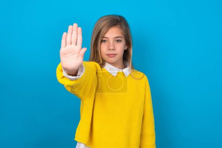 Foto de Chica adolescente caucásica con suéter amarillo sobre la pared azul haciendo señal de stop con la palma de la mano. Expresión de advertencia con gesto negativo y serio en la cara. - Imagen libre de derechos