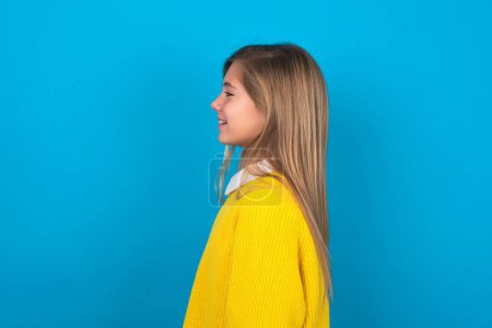 Photo for Caucasian teen girl wearing yellow sweater over blue wall looking to side, relax profile pose with natural face with confident smile. - Royalty Free Image